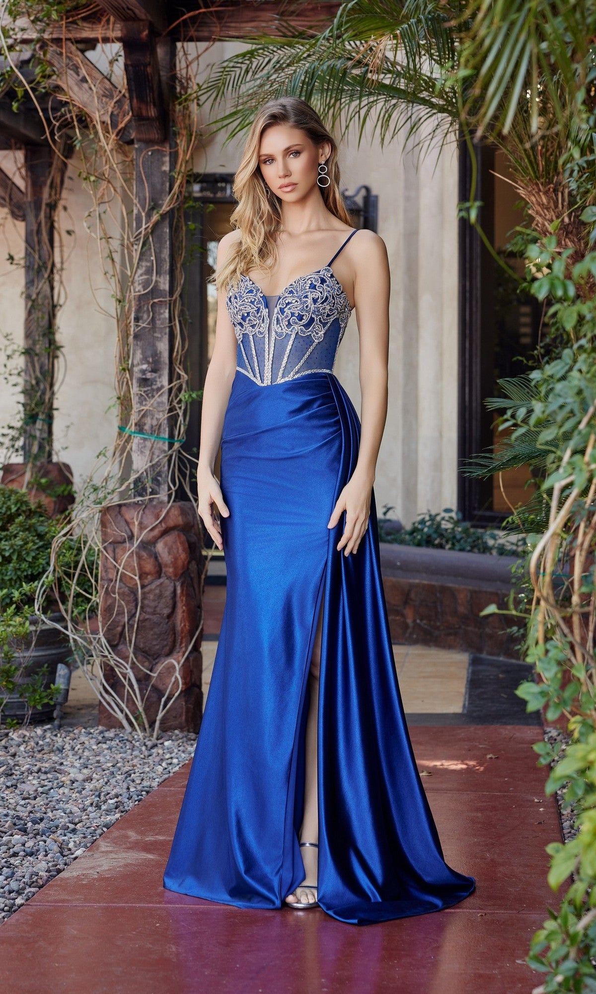 A woman poses outdoors on a red walkway surrounded by greenery, wearing the Nox Anabel G1788 long prom dress in navy blue, featuring a satin finish, beaded corset bodice, and a high slit.